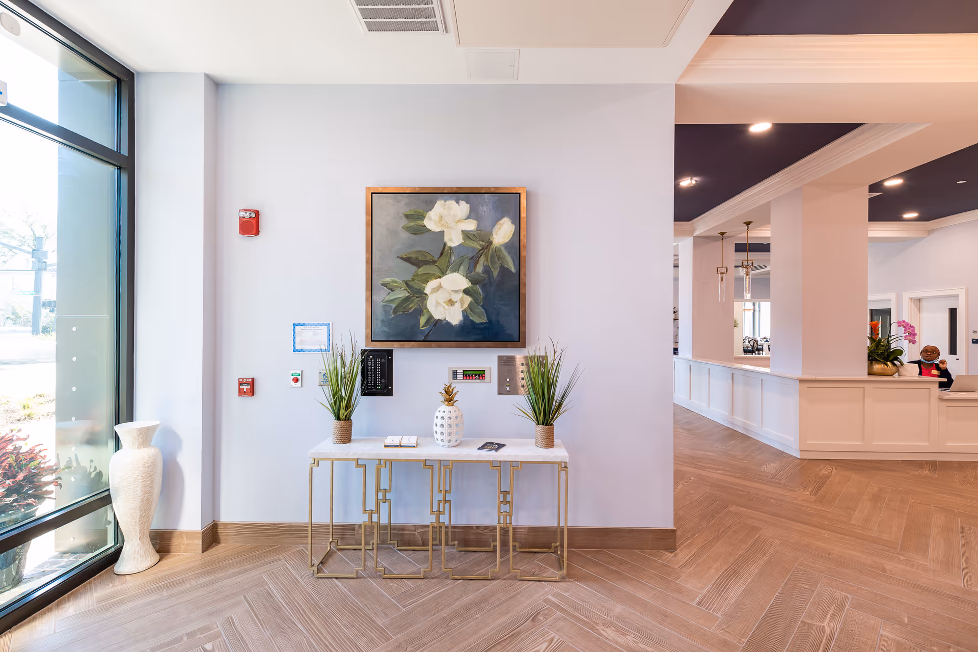 A bright and modern interior space with a large window on the left letting in natural light. A decorative console table with gold legs and a white top is placed against a light-colored wall, adorned with two small potted plants and a pineapple-shaped ornament. Above the table hangs a framed painting of white flowers with green leaves. To the right, there is a reception desk area with white paneling and a dark ceiling, featuring pendant lights and some decorative plants.