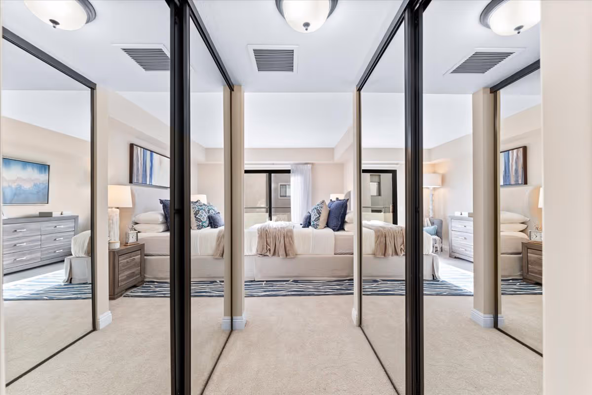 Mirrored closet doors reflecting a neatly made bedroom with a bed, nightstands, dressers, and lamps.