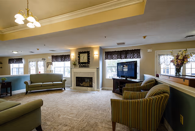 A spacious living room area in a senior living facility featuring a beige carpet, a fireplace with a clock above it, a flat-screen TV on a wooden stand, two striped armchairs, a green sofa, and windows with dark valances letting in natural light.