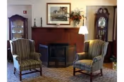 A cozy living room area with two striped armchairs facing each other in front of a wooden fireplace. The fireplace has a black metal screen and is decorated with a framed picture above it, two candlesticks, and a floral arrangement. To the left is a glass-fronted cabinet, and to the right is a tall grandfather clock. A floor lamp stands between the right armchair and the fireplace.