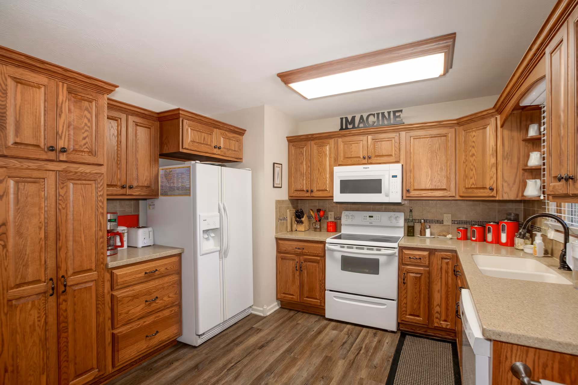 A kitchen with wooden cabinets, a white refrigerator, white microwave, and white stove. The countertops are beige, and there are red kitchen accessories including a coffee maker, canisters, and utensils. A sign that says 'IMAGINE' is displayed above the cabinets. The floor is wood-style laminate.