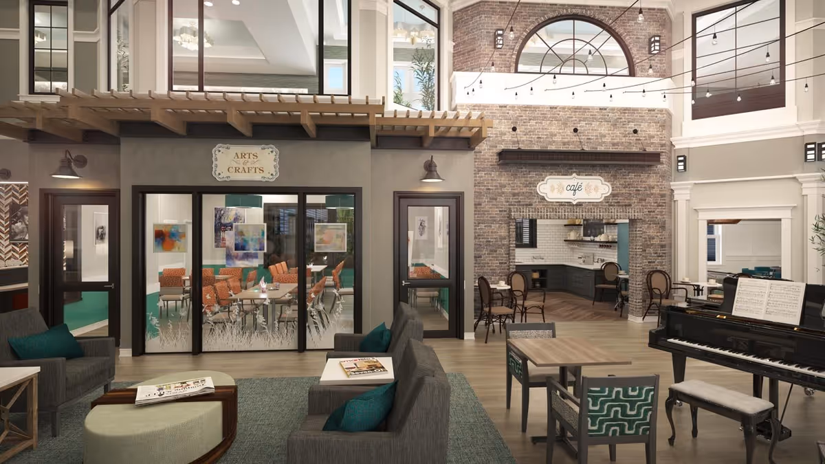 A spacious and well-lit common area in a senior living facility featuring a seating area with sofas and a coffee table, a grand piano with sheet music, a small dining table with chairs, and a café area with brick walls and a sign reading 'café'. There is also a glass-enclosed room labeled 'Arts & Crafts' with orange chairs and colorful artwork on the walls. The room has high ceilings with large windows and string lights hanging overhead.