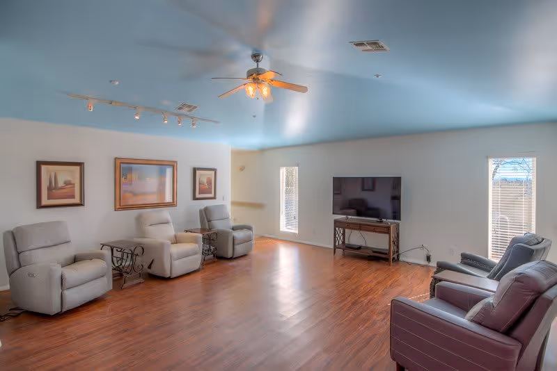 A spacious living room with wooden flooring, light blue ceiling, and white walls. The room features five recliner chairs arranged around the perimeter, three framed landscape paintings on one wall, a ceiling fan with lights, and a flat-screen TV on a stand positioned between two tall windows with blinds.