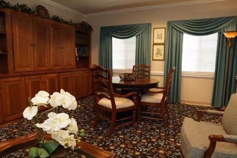 A cozy living room with a round wooden table surrounded by four cushioned wooden chairs, two windows with green drapes, a large wooden cabinet along one wall, a beige armchair, and a floral patterned carpet. A white orchid plant is visible in the foreground on a wooden coffee table.