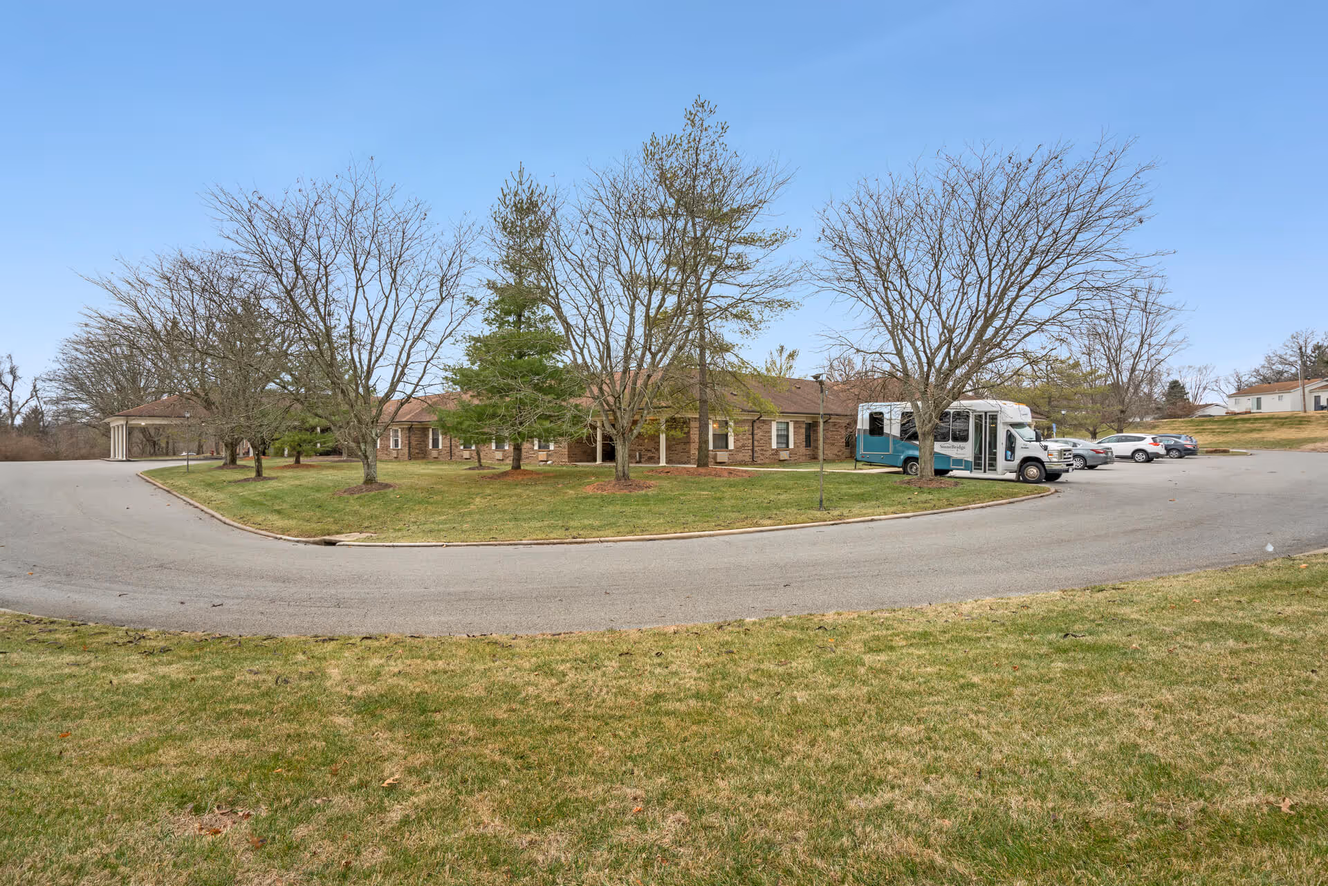 Exterior view of StoneBridge Senior Living facility showing a single-story brick building surrounded by leafless trees and a curved driveway with parked cars and a shuttle bus.