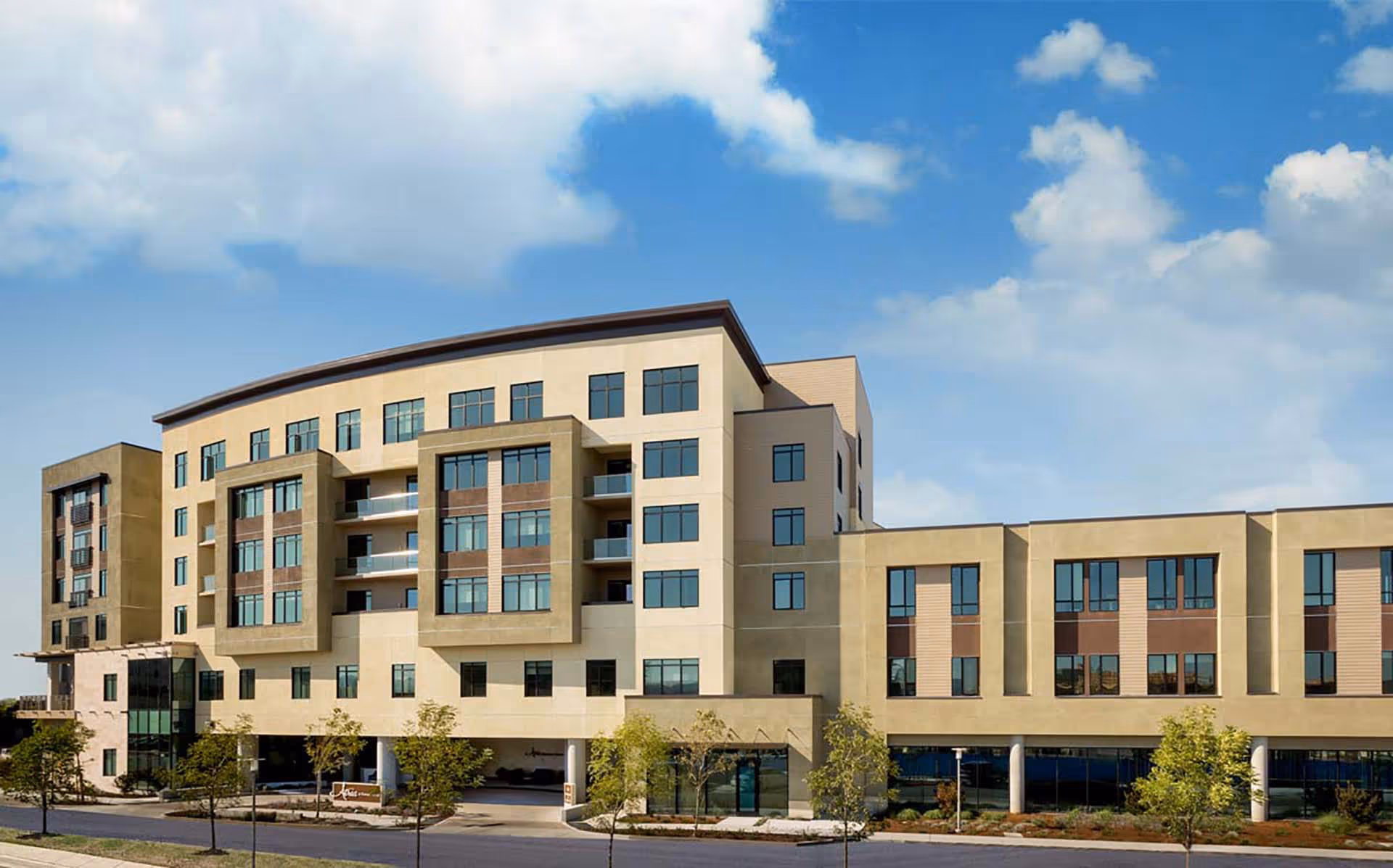 Exterior view of a modern multi-story senior living facility building with large windows, balconies, and landscaped surroundings under a partly cloudy blue sky.