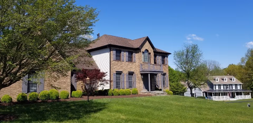 A large two-story brick and siding house with multiple windows and a brown roof, surrounded by green grass, bushes, and trees under a clear blue sky. Another house with a porch and dormer windows is visible in the background.