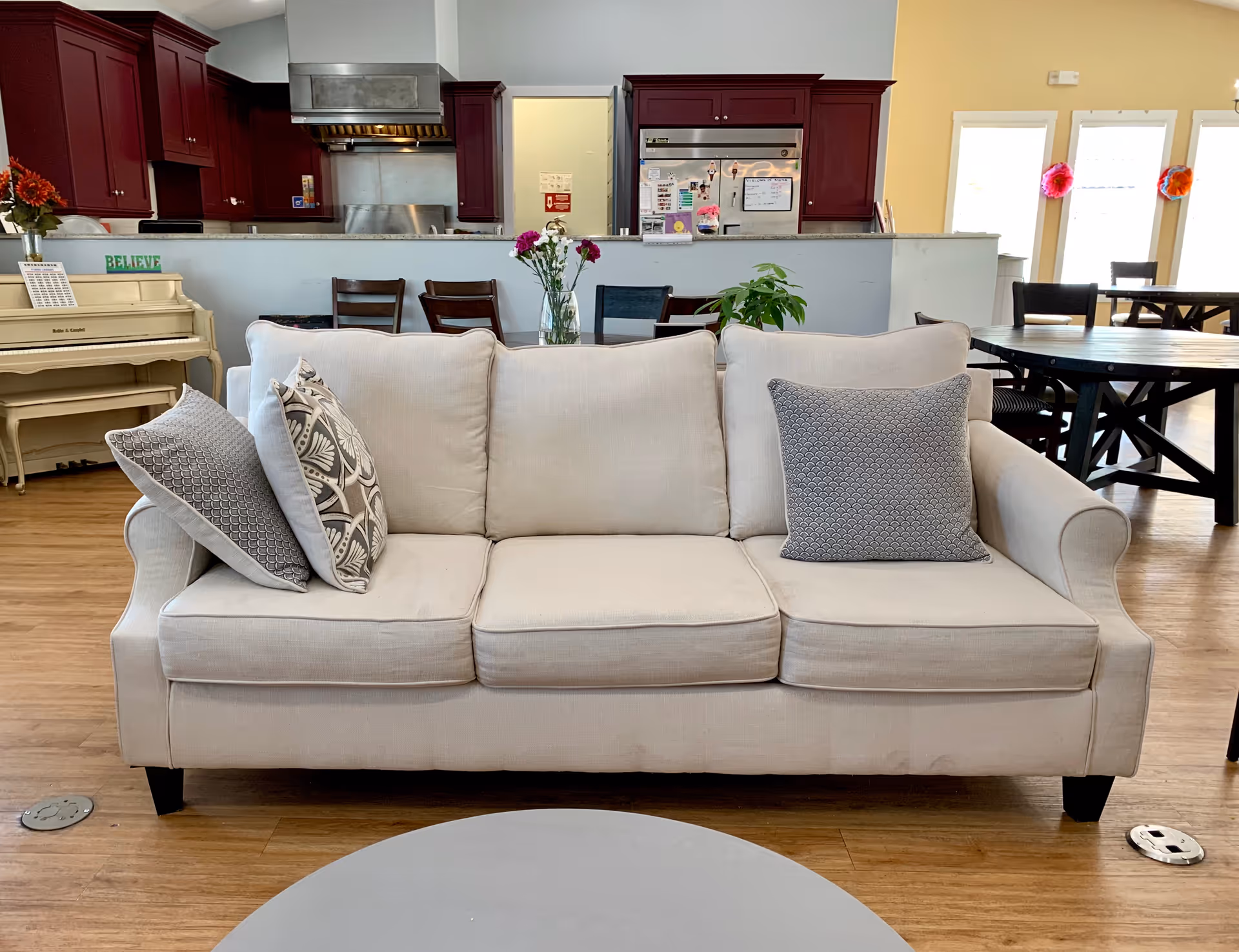 A beige sofa with three cushions in a common area. Behind the sofa is a dining table with chairs, a kitchen with dark red cabinets, and a white piano with a 'BELIEVE' sign on it. There are flowers in vases on the dining table and kitchen counter, and large windows letting in natural light.