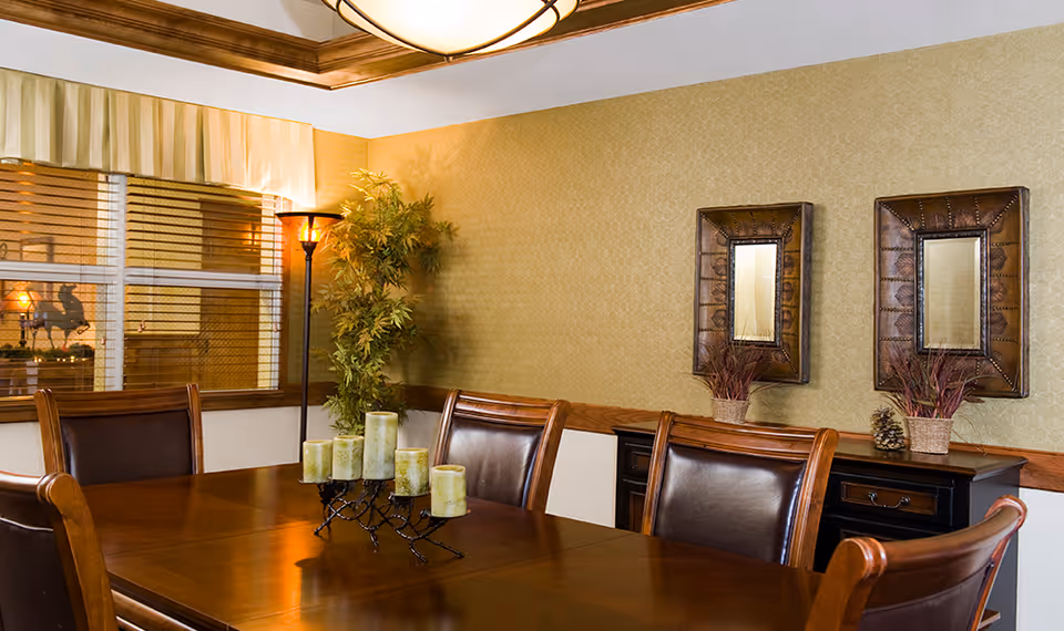 A warmly lit dining room with a wooden table and leather chairs, decorated with candles, mirrors, a floor lamp, and a potted plant.