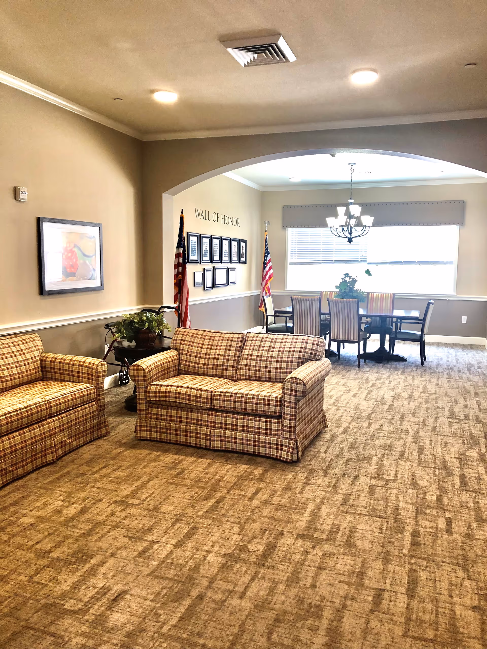 A cozy interior common area with two plaid sofas, a small table with a plant, and a dining table with six chairs under a chandelier. The wall in the background has a 'Wall of Honor' display with framed pictures and two American flags on either side.