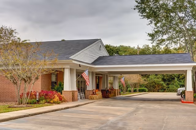 Exterior view of a single-story brick building with a covered driveway entrance. The entrance is decorated with American flags, hay bales, and autumn-themed decorations. Trees and shrubs surround the building under a cloudy sky.