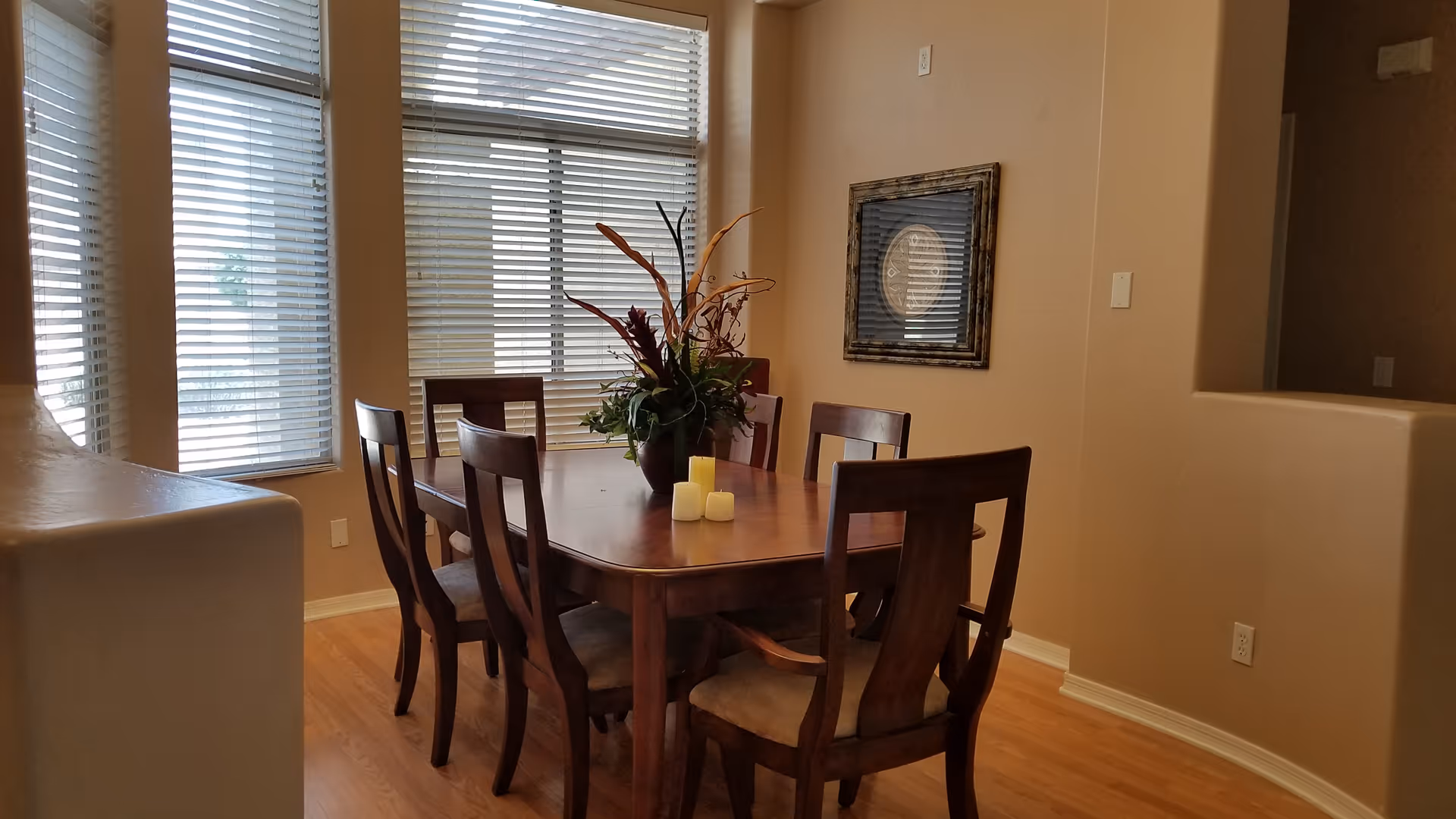 A dining room with a wooden rectangular table surrounded by six matching wooden chairs with cushioned seats. The table has a floral centerpiece and three candles. The room has light brown walls, hardwood flooring, and three large windows with white blinds letting in natural light. A framed decorative piece hangs on the wall.