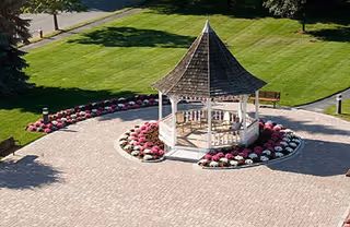 White wooden gazebo on a brick-paved circular plaza surrounded by pink and white flower beds and green lawn.