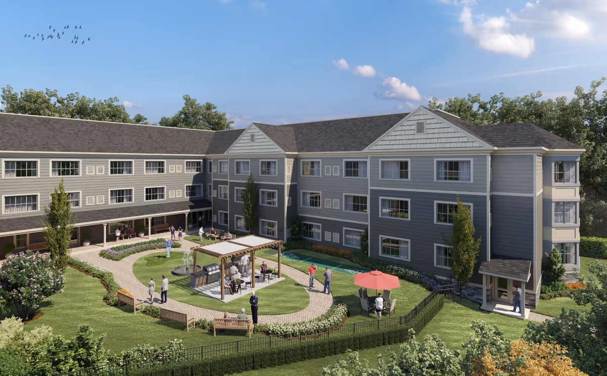 Exterior view of a three-story senior living facility building with gray siding and white trim. The building surrounds a landscaped courtyard with a walking path, benches, a pergola with seating, a small putting green, and several people enjoying the outdoor space. Trees and shrubs surround the area under a blue sky with some clouds and a flock of birds flying.