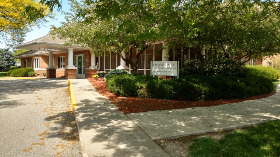 Exterior view of a single-story brick building with white columns at the entrance, surrounded by green trees and bushes, with a sidewalk leading to the entrance. A sign is visible among the bushes.