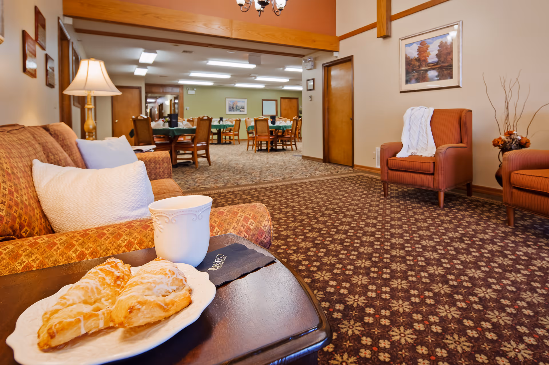 Cozy senior living common area with a sofa and armchairs, a table holding a cup and pastries, and a dining room visible in the background.