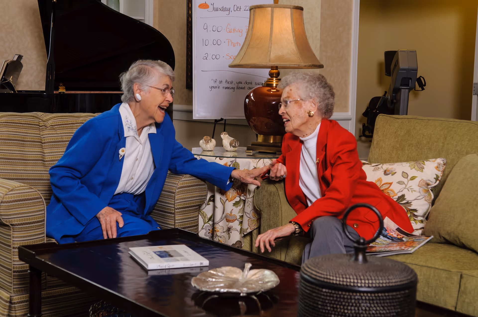 Two elderly women sitting and engaging in a lively conversation in a cozy living room setting. One woman is wearing a blue suit and the other a red jacket. They are seated on comfortable armchairs and a sofa with a coffee table in front of them. A lamp and a whiteboard with a schedule are visible in the background.