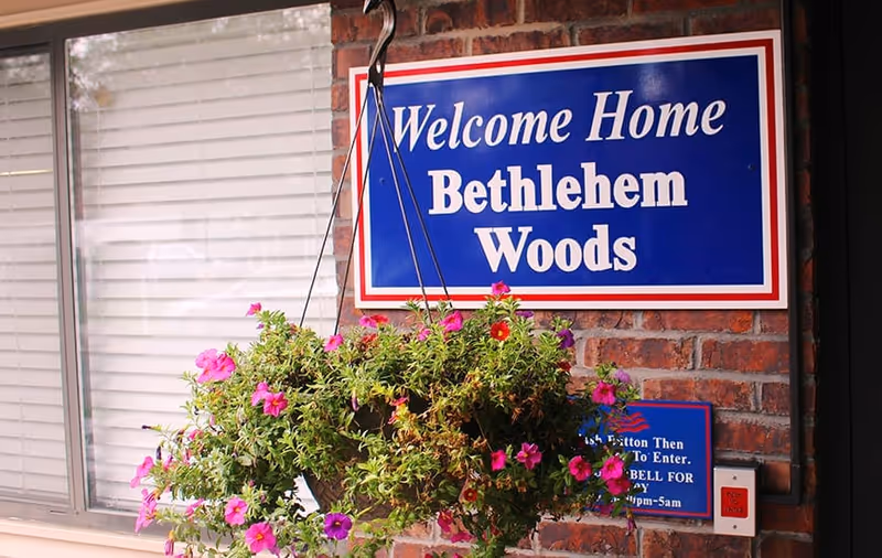 Blue 'Welcome Home Bethlehem Woods' sign on a brick wall above a hanging basket of pink flowers by a window.