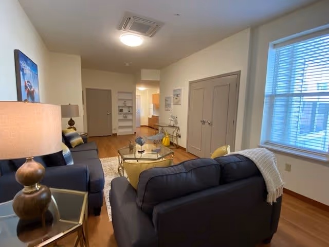 A cozy living room area in a senior living facility with two dark gray sofas, a glass coffee table with decorative items, a side table with a lamp, a window with blinds, and a view into a kitchen area in the background.