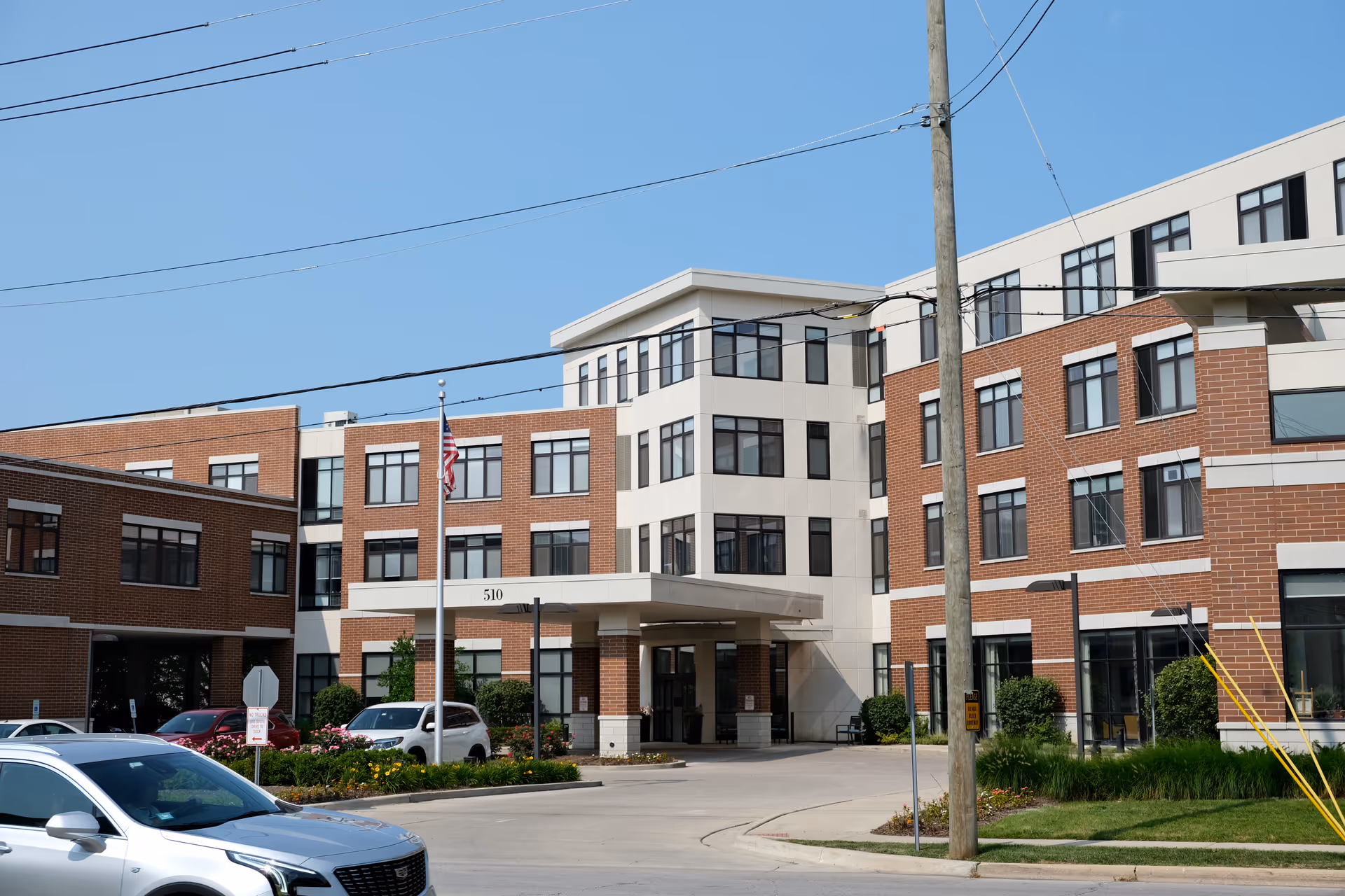 Exterior view of a multi-story senior living facility building with a brick and white facade, a covered entrance with the number 510, several parked cars, an American flag on a flagpole, and clear blue sky.