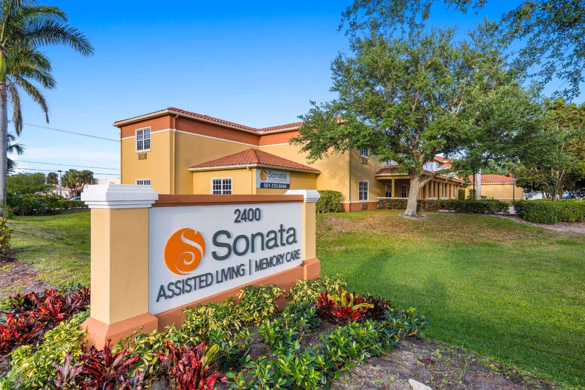 Exterior view of Sonata Boynton Beach assisted living and memory care facility with a large sign displaying the name and address 2400 Sonata. The building is a two-story structure with a tan and orange color scheme, surrounded by green grass, trees, and landscaping under a clear blue sky.