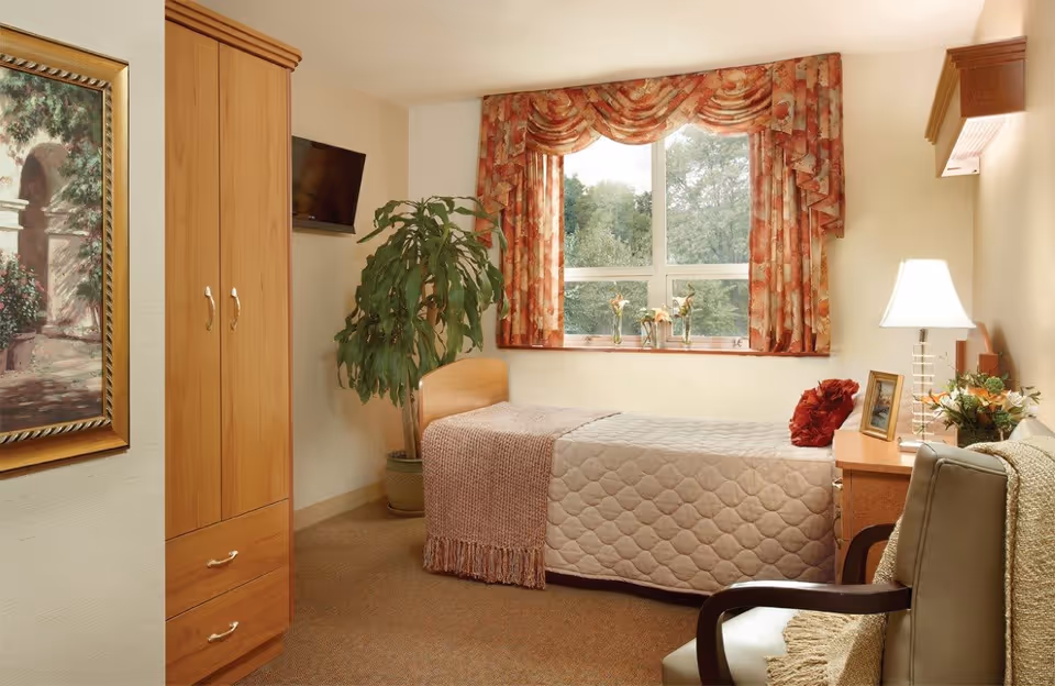 A cozy bedroom in Glenview Terrace featuring a single bed with a quilted bedspread and a knitted throw. The room has a wooden wardrobe, a small desk with a lamp, framed photo, and flower arrangement. A large window with floral curtains lets in natural light, and a potted plant stands beside the bed. A comfortable armchair with a throw blanket is also visible.