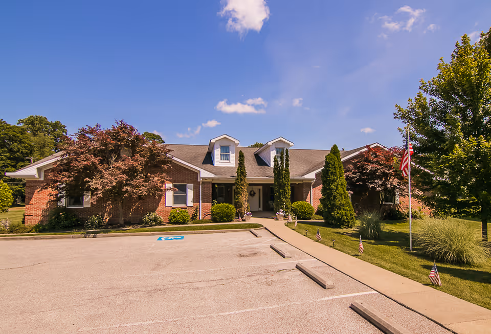 Front exterior of a single-story brick assisted living building with dormers, landscaped lawn, a parking area, and an American flag.
