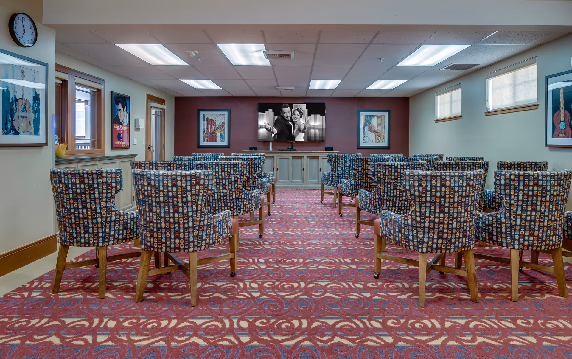 A small theater-style room with patterned chairs arranged in rows facing a large flat-screen TV mounted on a burgundy accent wall. The TV displays a black and white image of a man and woman. The room has a red carpet with a circular pattern, framed artwork on the walls, windows with shades, and a clock on the left wall.