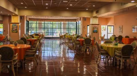 Bright dining hall with round tables, colorful cloths, and large windows in a senior facility.