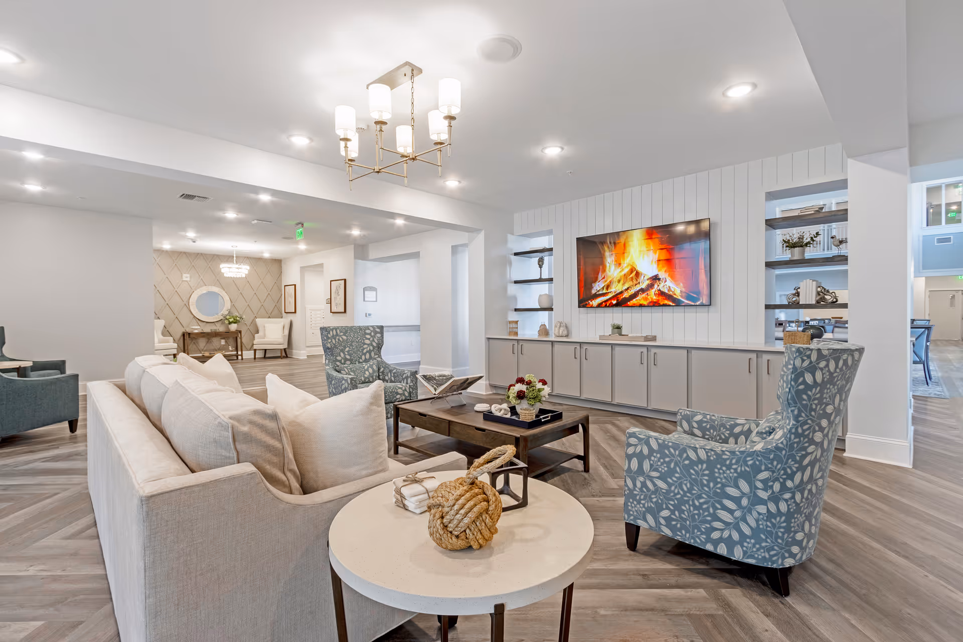 A bright and spacious living room area with a beige sofa, two patterned blue armchairs, a round side table with decorative items, and a rectangular coffee table with flowers and books. A large flat-screen TV mounted on a white paneled wall displays a fireplace scene. The room features wood flooring, built-in shelves with decorative items, and modern ceiling lights including a chandelier.