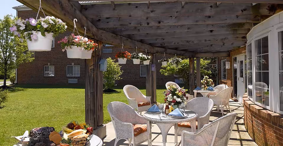 Outdoor patio area with white wicker chairs and tables set with blue napkins and floral centerpieces under a wooden pergola with hanging flower pots. The patio is adjacent to a brick building with large windows and a green lawn in the background.