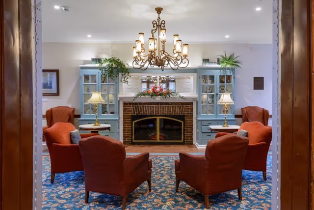 Cozy common room with four red upholstered armchairs arranged around a brick fireplace flanked by blue built-in cabinets, a chandelier overhead and patterned carpet.