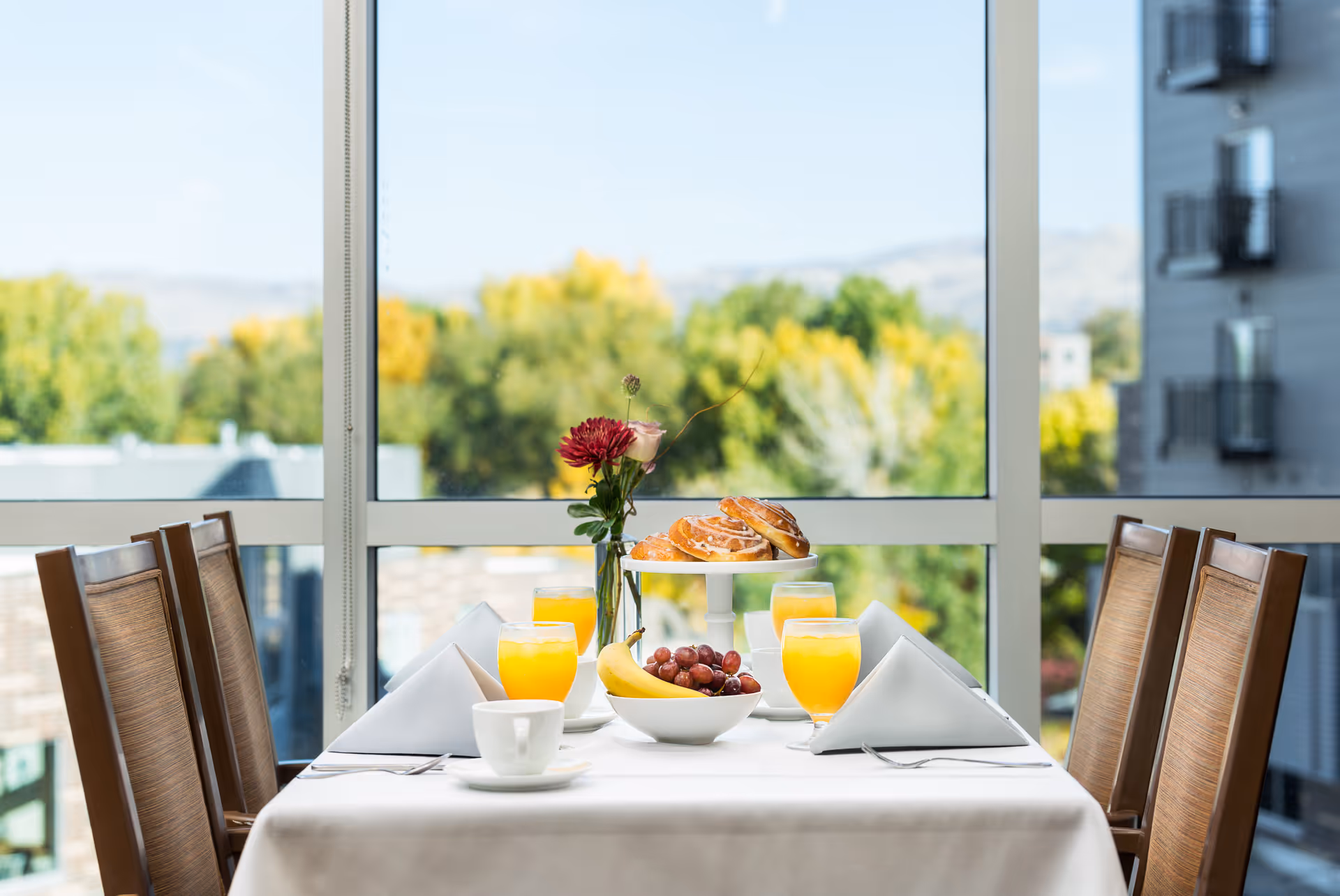A dining table set for four with white tablecloth, white napkins folded in triangles, four glasses of orange juice, a bowl of fruit including bananas and grapes, a tiered tray with pastries, and a vase with flowers. The table is positioned in front of large windows showing a blurred outdoor view with trees and a building.