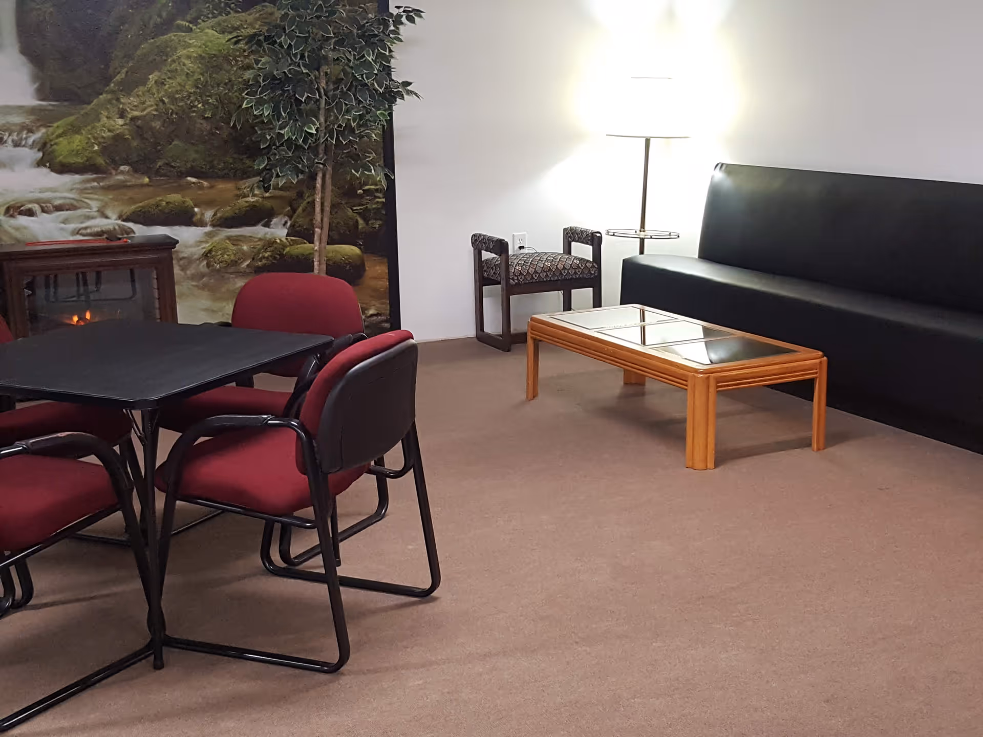 Community lounge with maroon chairs around a table, a black sofa, a wooden coffee table, and a nature mural on the wall.