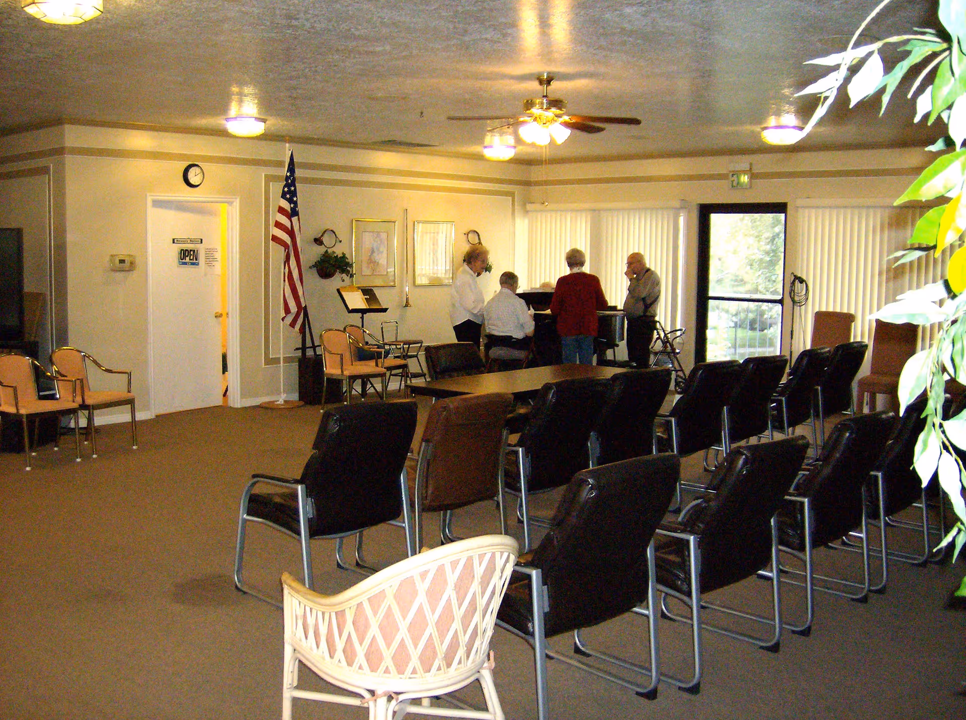 A community room with rows of chairs facing a piano where four elderly people are gathered. The room has beige walls, carpeted floor, an American flag, framed pictures, and large windows with vertical blinds. There is a ceiling fan with lights and some plants visible on the right side.