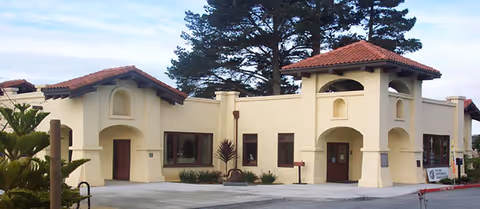 Cream-colored Mediterranean-style building with red tile roofs, arched windows and entryways in front of tall trees under a clear sky.