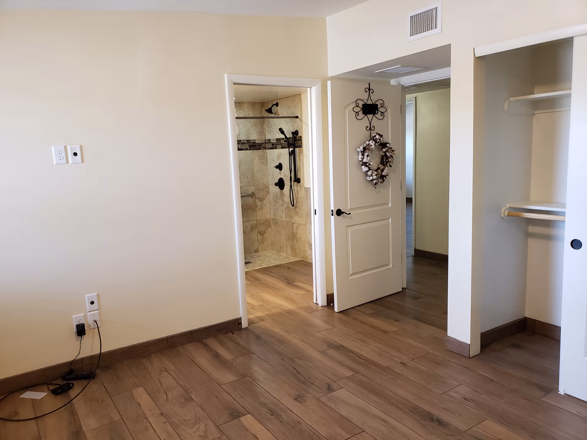 Empty room with wooden flooring, an open door leading to a tiled shower area, and an open closet with sliding doors. The door has a decorative wreath hanging on it.
