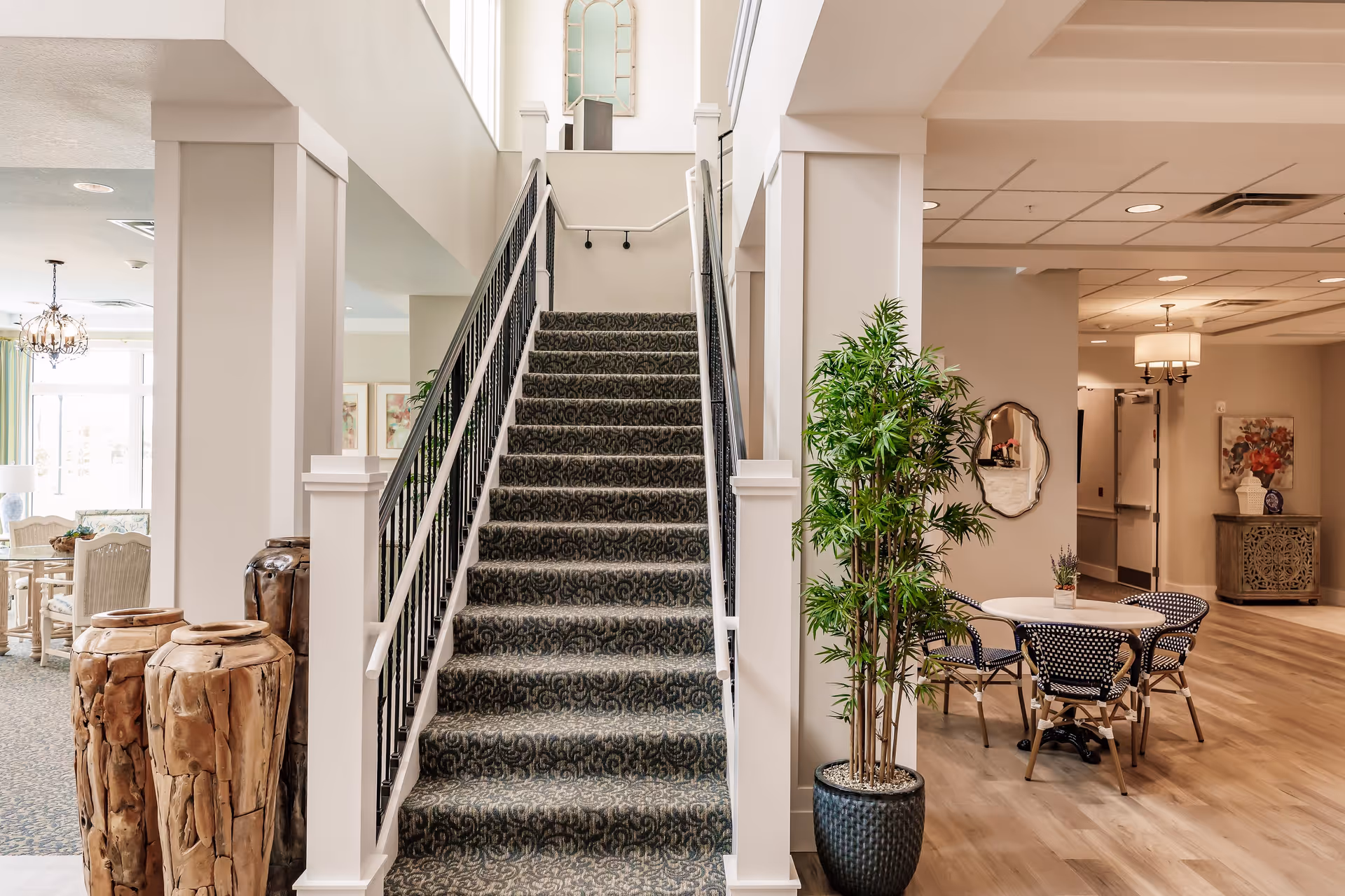 Interior view of a senior living facility featuring a carpeted staircase with black railings in the center. To the right, there is a round table with four black and white patterned chairs and a tall potted plant. The area has light-colored walls and wood flooring, with decorative elements including a mirror, a floral painting, and a chandelier in the background.