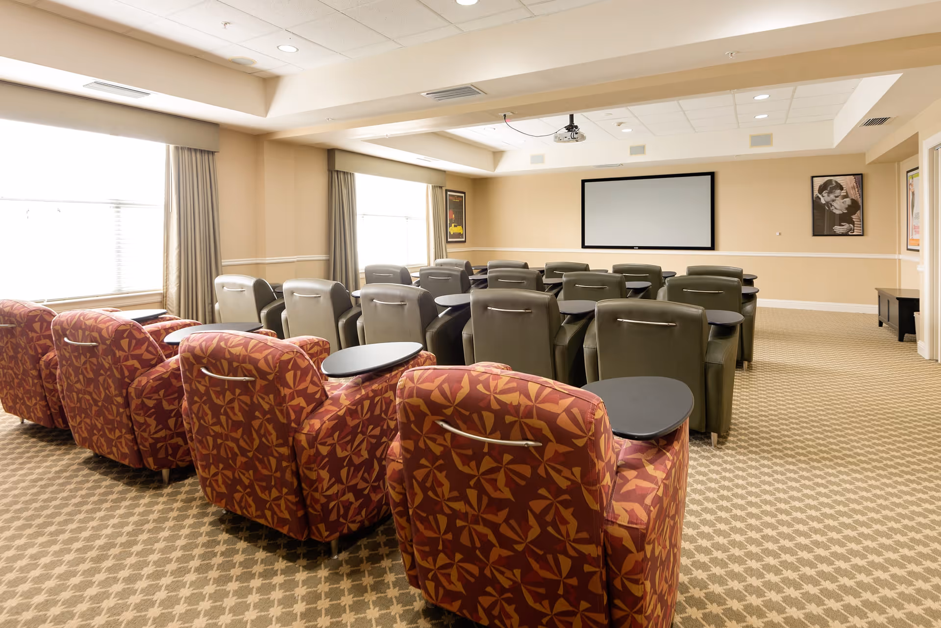 A senior living facility media room with rows of comfortable chairs facing a large blank projection screen. The chairs in the front row are patterned in red and orange, while the rest are gray. The room has beige walls, carpeted floor, and windows with curtains letting in natural light.