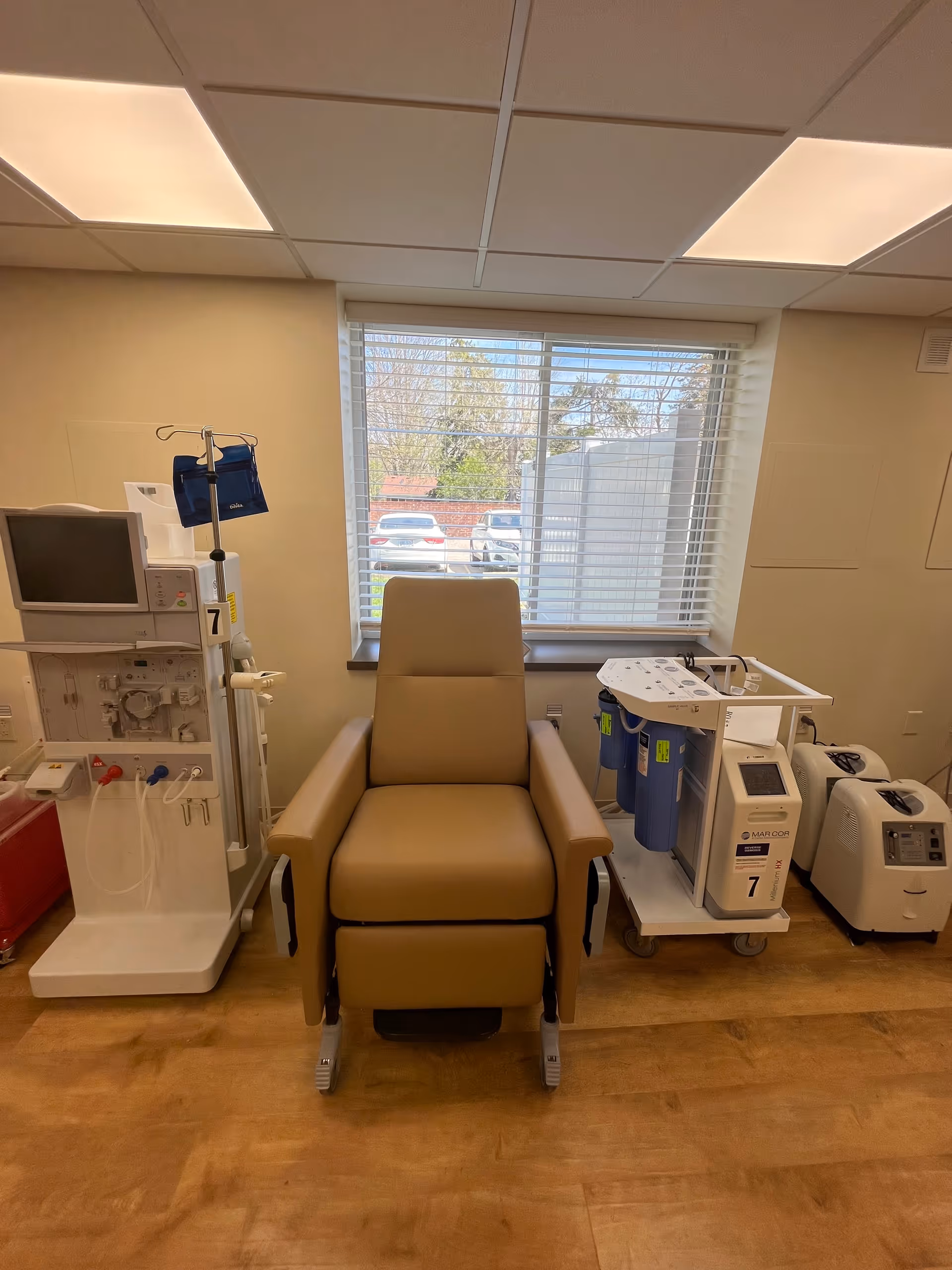 A medical treatment room with a beige recliner chair positioned in front of a window with blinds. To the left of the chair is a dialysis machine with tubes and a monitor, and to the right are medical filtration devices and oxygen concentrators on a wooden floor.