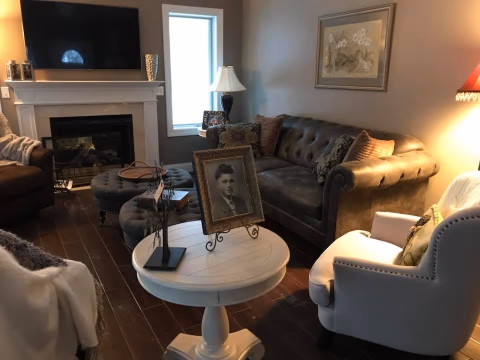 A cozy living room with a dark tufted sofa adorned with pillows, a white armchair with a decorative pillow, and a round white wooden table in the center holding a framed black-and-white portrait and a small decorative tree. The room features a fireplace with a flat-screen TV mounted above it, a window letting in natural light, a table lamp on a side table, and a framed floral artwork on the wall.