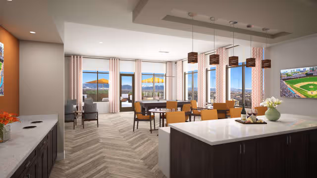 A spacious and modern common area in a senior living facility featuring a large kitchen island with a white countertop and dark wood cabinets. The room has multiple seating areas with tables and chairs, large windows with pink patterned curtains letting in natural light, and a wall-mounted TV displaying a baseball game. Outside the windows, yellow umbrellas are visible on a patio with a scenic view of mountains in the distance.