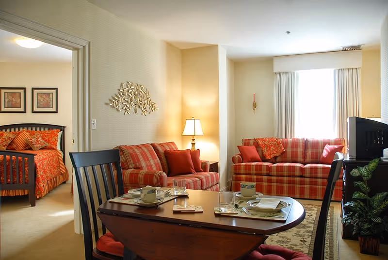 A cozy living room with two red plaid sofas, a wooden dining table set with cups and plates, a lamp on a side table, and a window with white curtains. In the background, a bedroom with a bed covered in red and orange bedding is visible through an open doorway.