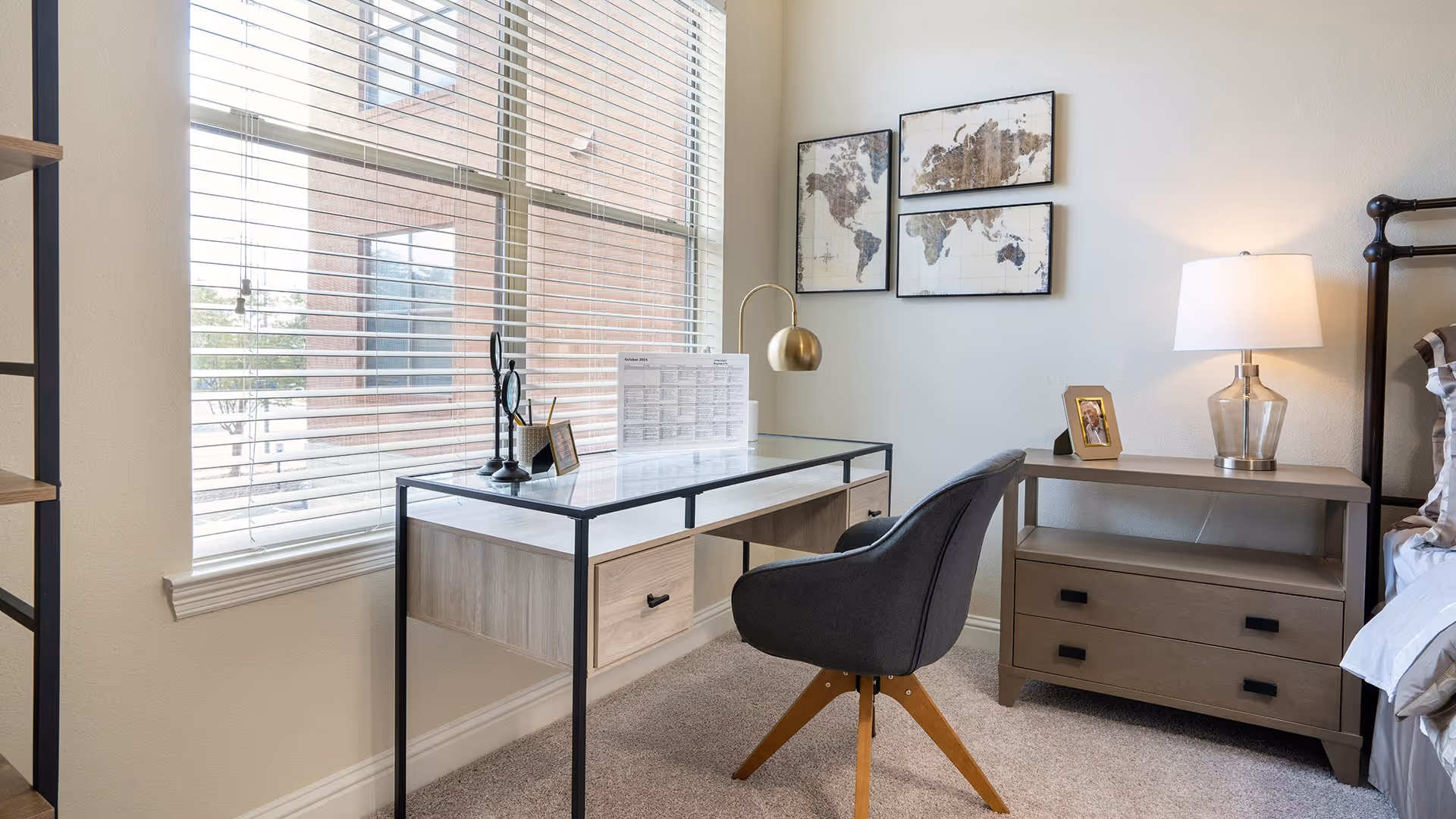 Bright bedroom corner with a glass-top desk by a large window, a gray swivel chair, nightstand with a lamp, and world map wall art.