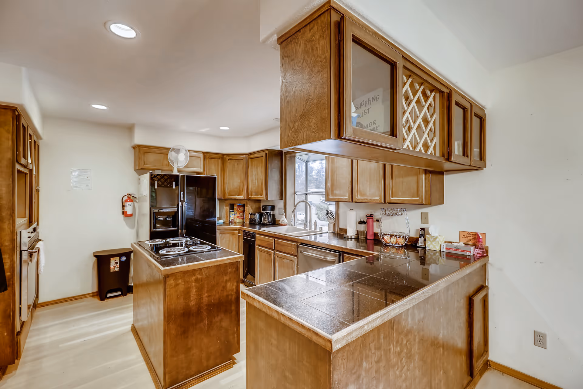 A bright kitchen with wooden cabinets and a black countertop island featuring an electric stove. The kitchen includes a black refrigerator, a coffee maker, a dishwasher, and a window above the sink letting in natural light. Various kitchen items such as a paper towel holder, a basket of eggs, and a pink water bottle are visible on the counters.