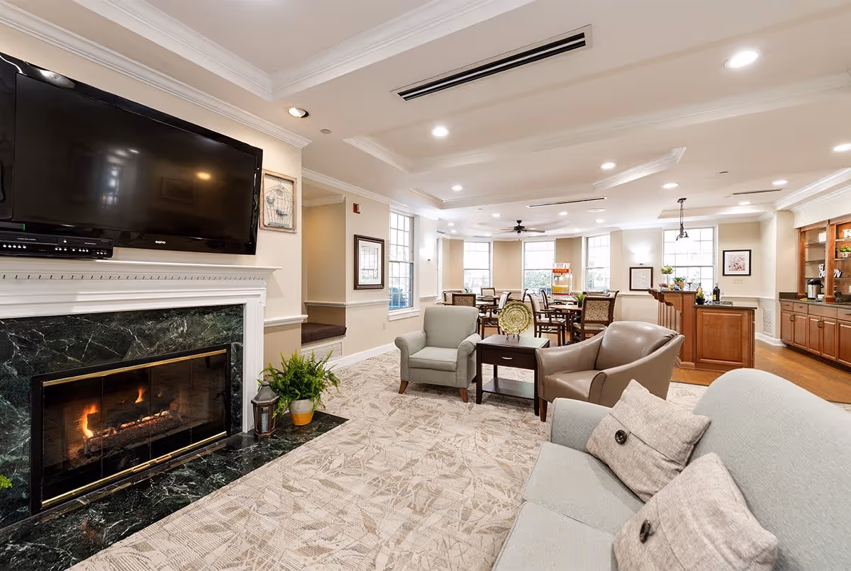 A spacious and well-lit living room area in a senior living facility featuring a large flat-screen TV mounted above a green marble fireplace with a fire burning. The room has comfortable seating including a sofa with pillows, armchairs, and a small table. In the background, there is a dining area with tables and chairs, large windows letting in natural light, and a wooden cabinet with a countertop and decor items.