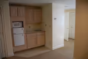 Small kitchenette area with light wood cabinets, a built-in microwave, and a small refrigerator. The kitchenette is adjacent to a hallway leading to a white door, with beige walls and carpeted floor.