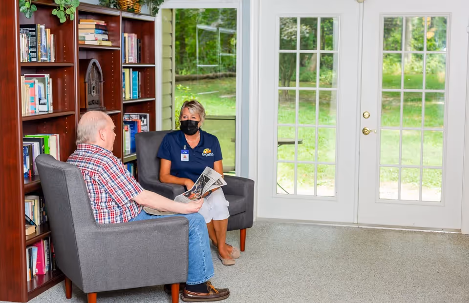 An elderly man and a woman wearing a mask and a name tag sit in gray armchairs facing each other in a room with bookshelves and large glass doors showing a green outdoor area.