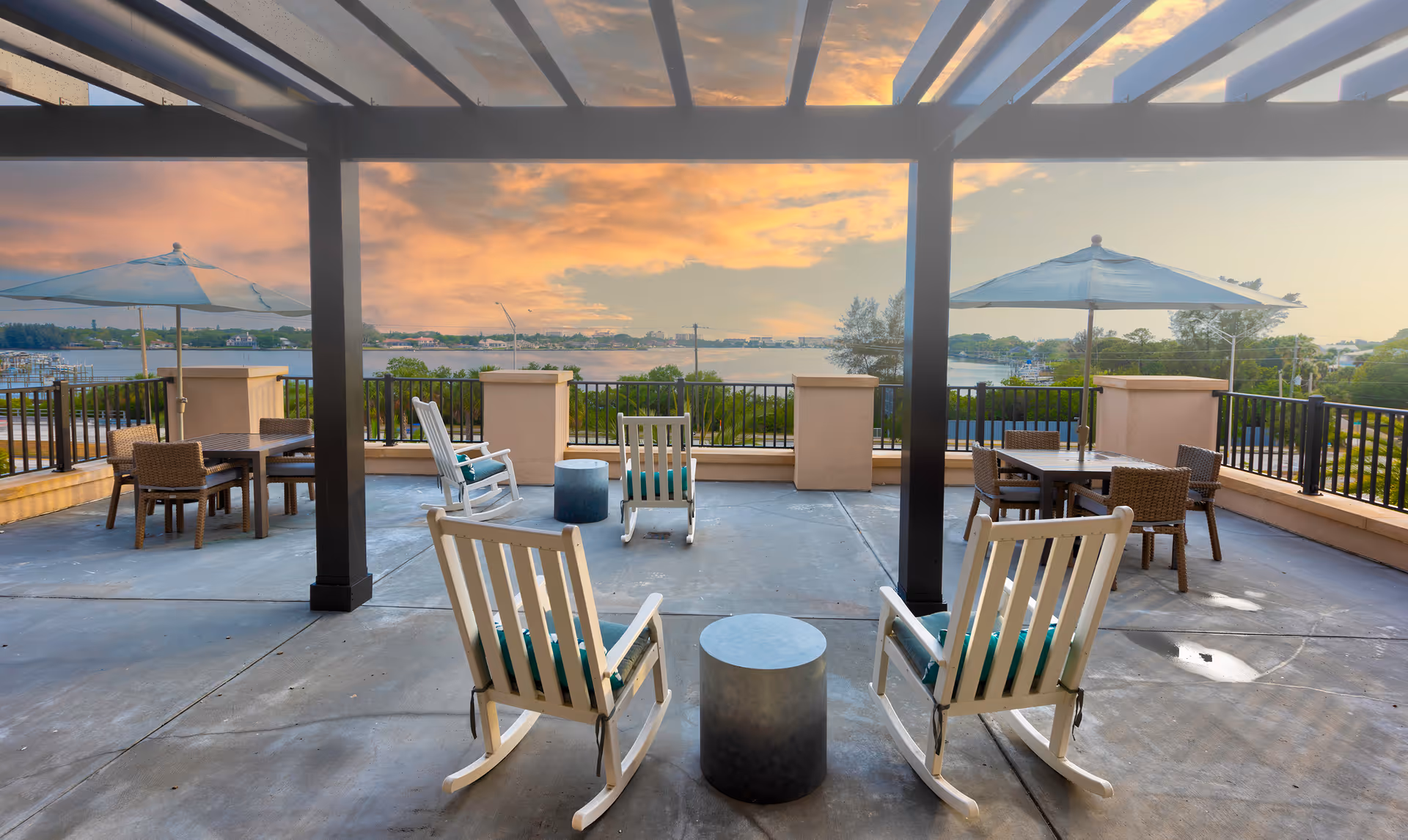 Outdoor patio area with white rocking chairs and small round tables under a pergola, overlooking a scenic waterfront view during sunset. There are also tables with umbrellas and wicker chairs on the patio.