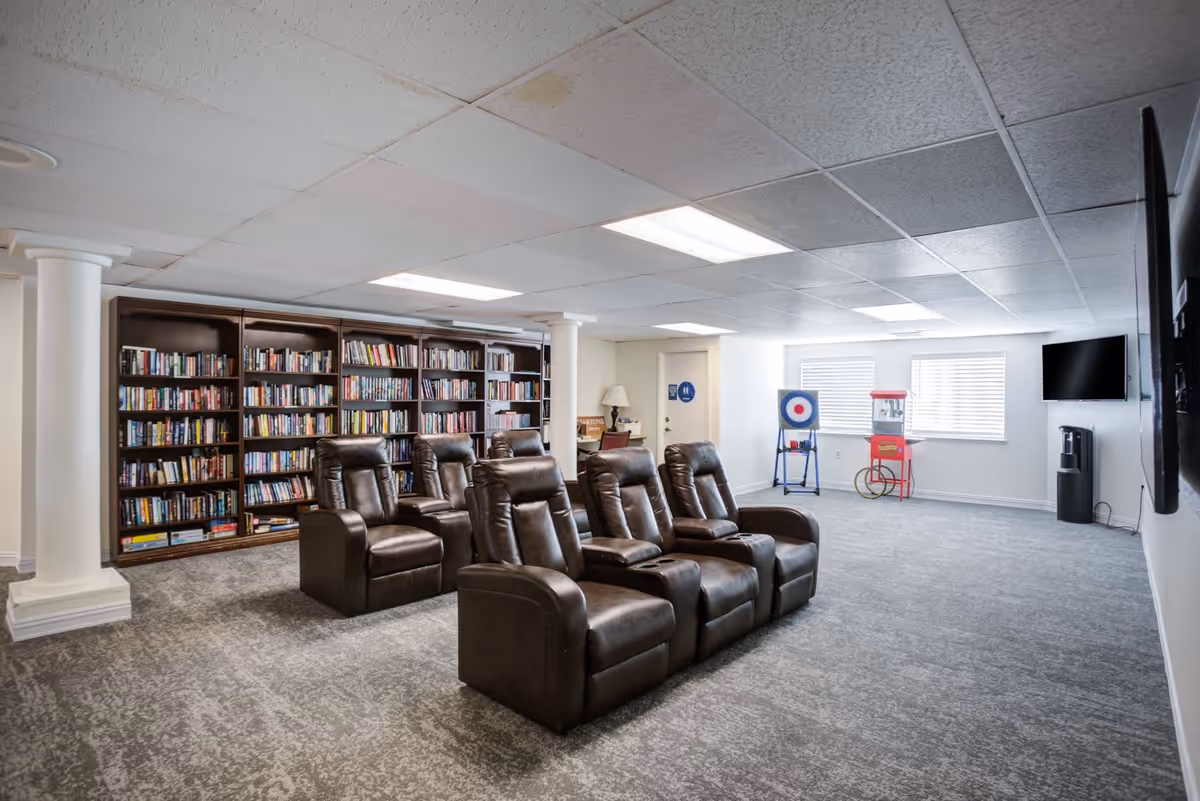 A spacious room with six brown leather recliner chairs arranged in two rows facing a wall-mounted flat screen TV. Behind the chairs is a large bookshelf filled with books. The room has carpeted flooring, white walls, and a drop ceiling with fluorescent lighting. In the background, there is a popcorn machine and a target game near two windows with blinds, and a water cooler is positioned near the TV.